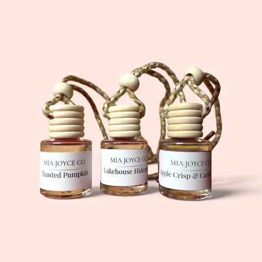 Three small glass reed diffuser bottles with white labels from MIA JOYCE CO. on a solid pale pink background.