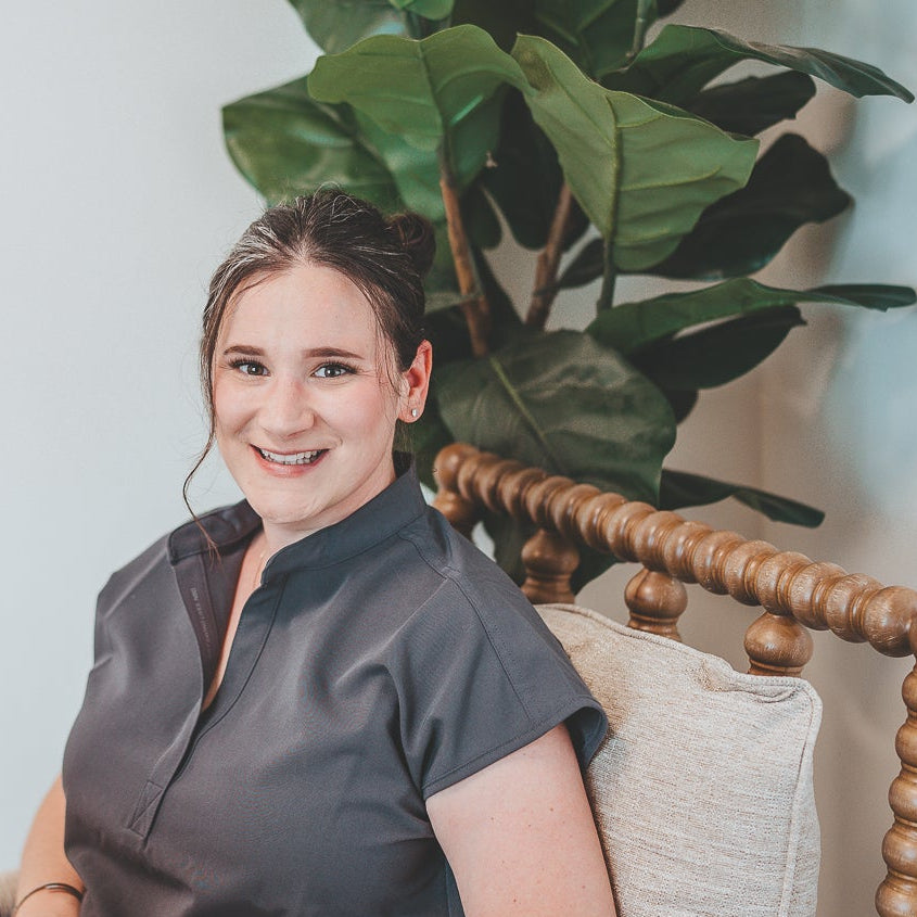A woman wearing a dark gray short-sleeve top and dark pants sits in a wooden armchair with a laptop on her lap, positioned in front of a large green fiddle-leaf fig plant against a white wall.