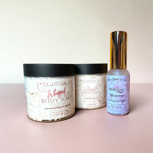 Jars of body scrub and body soap and a bottle of perfume on a pink surface with a white background